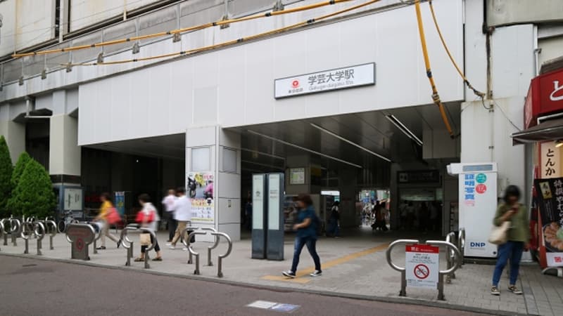 学芸大学駅の高架下にある白い外壁の入り口。駅名の看板の下を、数人の歩行者が行き交っている様子。