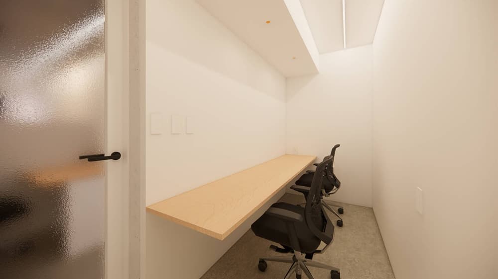 すりガラスのドアがある細長い個室。白い壁に固定された明るい木目のカウンターデスクと、黒い高機能オフィスチェアが2脚配置されている。
